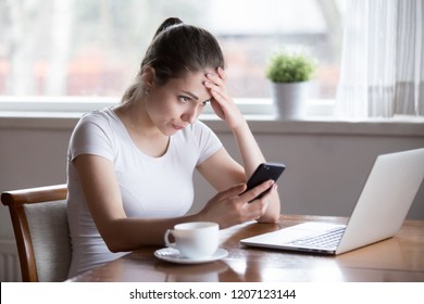 Annoyed Millennial Woman Sitting Alone In Kitchen At Table At Home Holding Smartphone Gadget Looking At Computer Screen Reading Message Notification Feels Irritated And Upset Have Problems With Device