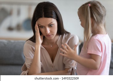 Annoyed Mom And Restless Little Daughter At Home, Mother Sitting On Couch Touch Head Or Temples Feels Headache Due To Noisy Kid Standing Near Her, Tired Parent Of Difficult Hyperactive Child Concept