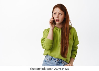 Annoyed teen girl with red hair talks on phone, roll eyes bothered and irritated by boring conversation, standing displeased against white background