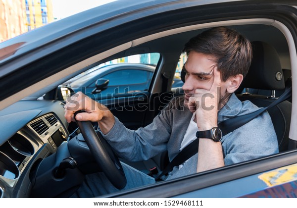 Annoyed tired young man is riding driving car.\
Businessman is late for meeting. Driver brunette in grey suit stuck\
in traffic jam. Stressful situations on roads and fast rhythm in\
modern city.