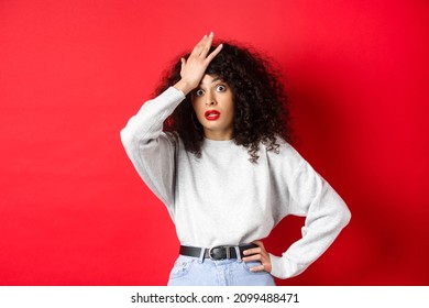 Annoyed and tired young woman slap forehead, making facepalm and stare at camera shocked, standing bothered against red background