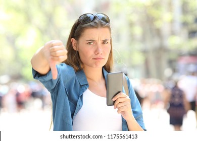 Annoyed Woman Holding A Smart Phone Looking At Camera With Thumbs Down In The Street