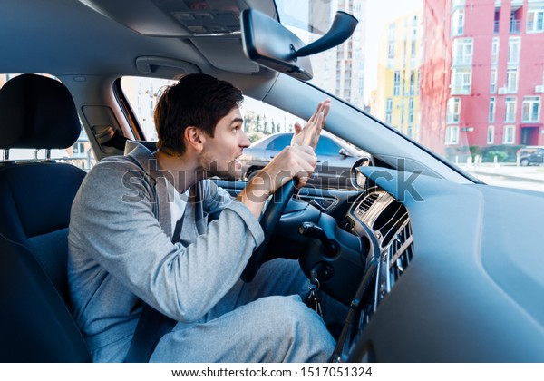 Annoyed young businessman is driving car. Man in\
gray suit is swearing with other road users. Brunette male is\
violating rules and laws. Stressful emergency situations on routes\
in modern city.