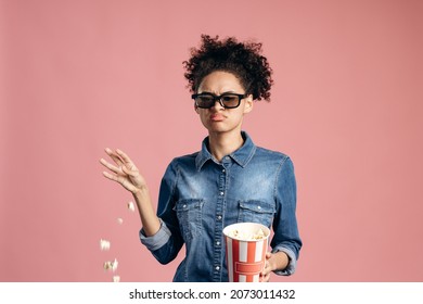 Annoyed young female in 3d imax glasses watching movie film, throwing popcorn, being disappointed with the movie. Indoor studio shot isolated on pink background 