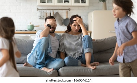 Annoyed Young Parents Sit On Couch In Kitchen Tired From Loud Two Little Children Running Playing, Anxious Mom And Dad Relax On Sofa At Home Exhausted From Active Ill-behaved Small Preschooler Kids