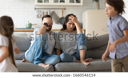 Annoyed young parents sit on couch in kitchen tired from loud two little children running playing, anxious mom and dad relax on sofa at home exhausted from active ill-behaved small preschooler kids