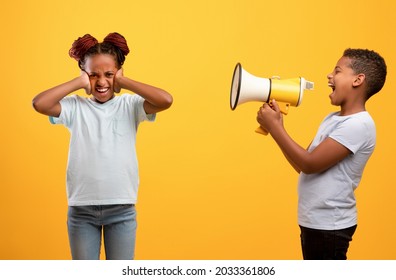 Annoying black boy screaming towards girl his sister through loudspeaker, yellow studio background. African american kids siblings fighting, teen girl covering her ears while naughty boy yelling