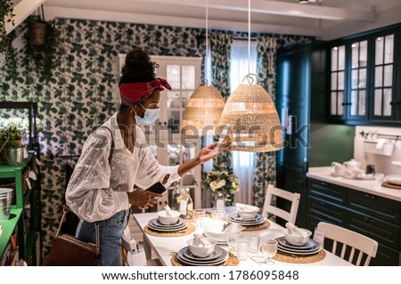 Anonymous African American female customer in casual clothes and sterile mask looking at price tag near lamp and in furniture shop during COVID 19 pandemic