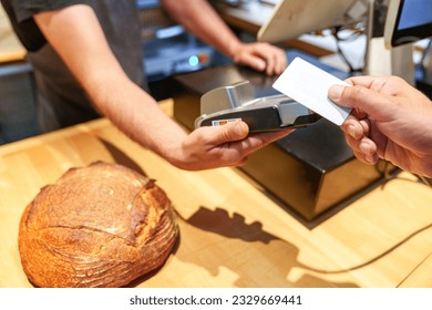 An anonymous bakery worker's hands use a card reader to accept payment from a customer for a loaf of bread.