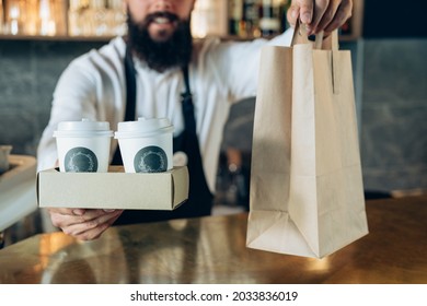 An Anonymous Barista Holding A Take Away Order.

Close Up Photo Of An Unrecognizable Male Hands Holding And Serving Disposable Tray With Two Cups Of Take Out Coffee And Food In Paper Bag To Customer.