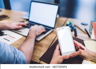 Anonymous blurred man using laptop with empty screen and female colleague texting on smartphone while sitting together at desk in office