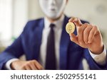 Anonymous business man in a suit and face mask holds a golden symbolic bitcoin in his hand, offers a safe exchange transaction and guarantees privacy. Selective focus close up. Crypto currency concept