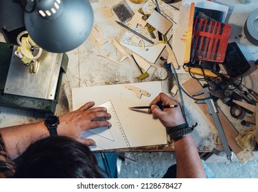 An Anonymous Carpenter Working On New Project At His Workshop. 
Table Top View Of Unrecognizable Man Drawing New Parts Using A Pen And Ruler On A Paper While Sitting At His Work Bench.