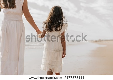 Anonymous child in white dress with long hair holding hand of mom while spending cloudy weekend day on beach near sea