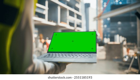 Anonymous Construction Manager Holding a Laptop with Green Screen Mock Up Template. Specialist Using Computer to Oversee the Project at a Construction Site - Powered by Shutterstock