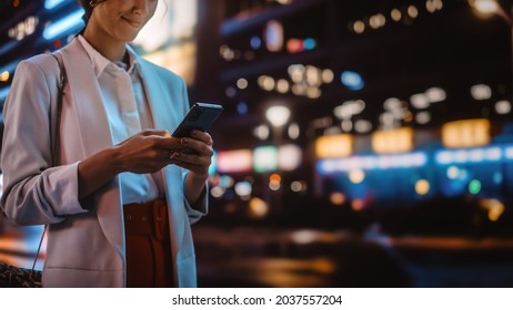 Anonymous Female Using Smartphone Walking Through Night City Street Full Of Neon Light. Woman Holding Mobile Phone, Posting Social Media, Online Shopping, Texting.