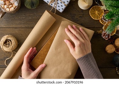 Anonymous female wrapping christmas gift in craft paper on wooden table. New Year decorations. Wintertime handcraft. Preparation for celebration. Hand-on activity concept