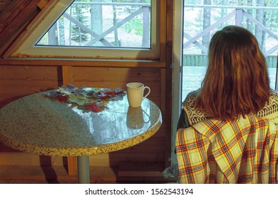 Anonymous Girl Sitting In The Attic Of A Rural House In Autumn. Woman Looking Outside The Window Near A Round Table With A Mug And Fall Foliage.