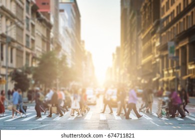 Anonymous Group Of People Walking Across A Pedestrian Crosswalk On A New York City Street With A Glowing Sunset Light Shining In The Background