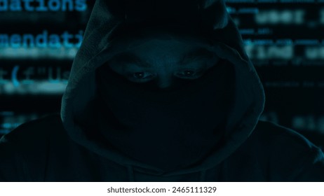 An anonymous hacker. Hooded figure with a masked face, program code in the background in a dark setting. - Powered by Shutterstock