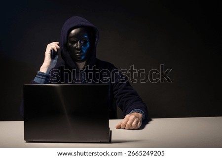 Anonymous hacker in mask programmer uses a laptop and talking on the phone to hack the system in the dark. The concept of cybercrime and hacking database