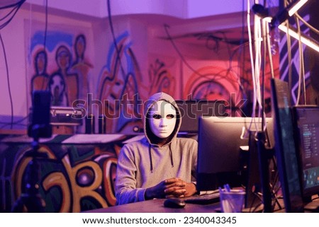 Anonymous hacker streaming threat message while doing illegal activities on computer. Internet thief recording online scam video on smartphone while breaking law in abandoned warehouse