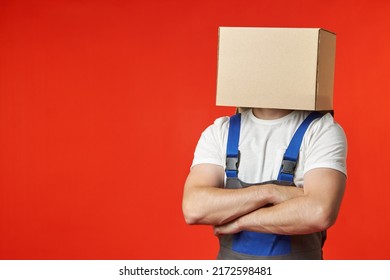 Anonymous Loader Confidently Stands In A Work Suit With Straps On A Red Background With A Box On His Head. A Professional Male Loader Thinks Only About The Safety And Security Of The Transported Items