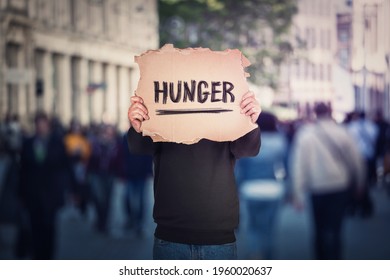 Anonymous man covers his head using a torn cardboard banner with hunger text message. Incognito person on a crowded street as protest demonstration against global famine. Food supply issue