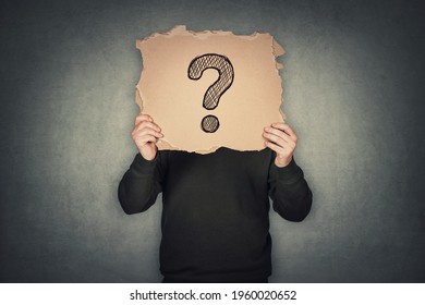 Anonymous man covers his head using a torn cardboard sheet, like a mask, with drawn question mark. Incognito person hidden, isolated on gray wall background. Introvert people hiding identity