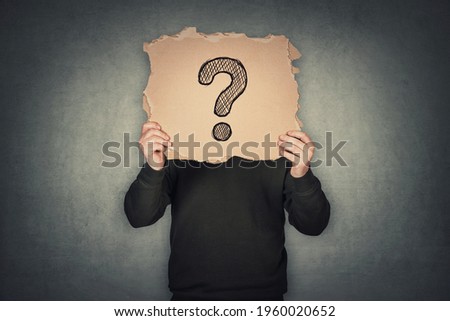 Anonymous man covers his head using a torn cardboard sheet, like a mask, with drawn question mark. Incognito person hidden, isolated on gray wall background. Introvert people hiding identity