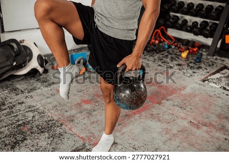 Anonymous man performs Single Leg Balance With Weight, using a kettlebell. Hardcore gym setting.