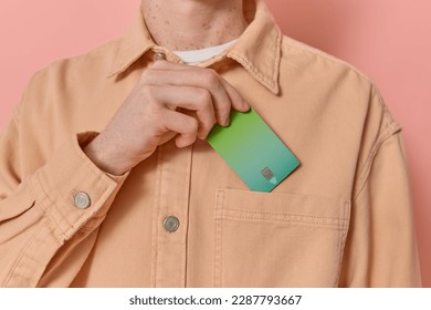 Anonymous man puts his green credit card in his pocket after payment on pink background copy space. Male consumer makes use of contactless payment methods like NFC. Modern banking, finance concept. 