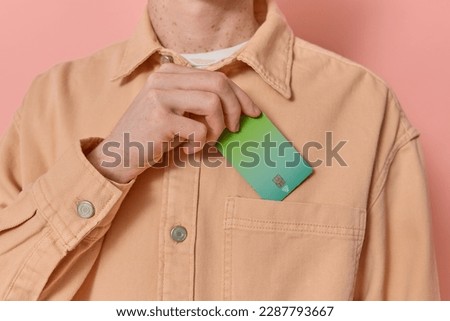 Anonymous man puts his green credit card in his pocket after payment on pink background copy space. Male consumer makes use of contactless payment methods like NFC. Modern banking, finance concept. 