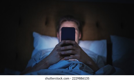 Anonymous Man Uses Smartphone in Bed at Home at Night. Handsome Guy Browsing Social Media, Reading News, Doing Online Shopping Late at Night. Focus on Hand Holding Mobile Phone Covering Face
