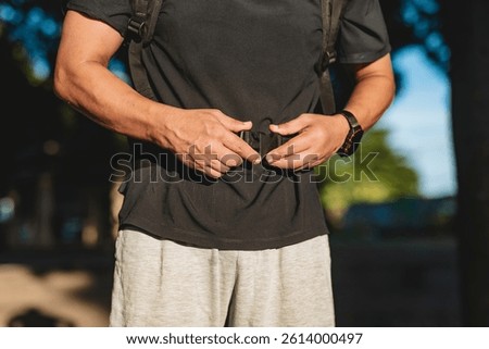 Anonymous middle aged man preparing for a ruck walk by securing the waist strap of a weighted backpack in a sunny park. Closeup shot.
