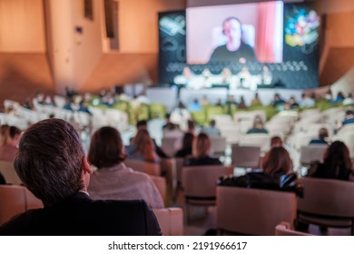 Anonymous People Sitting On Chairs And Listening To Woman Giving Speech From Large Screen During Business Conference In Spacious Auditorium