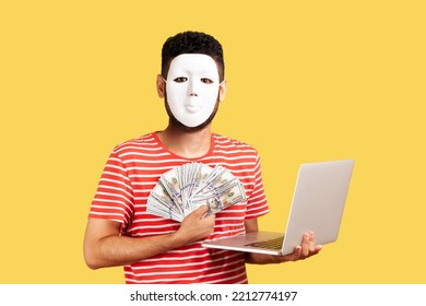 Anonymous Rich Hacker Or Programmer Man Hiding Face With White Mask, Hiding Personality, Conspiracy And Privacy, Working On Laptop And Showing Money. Indoor Studio Shot Isolated On Yellow Background.