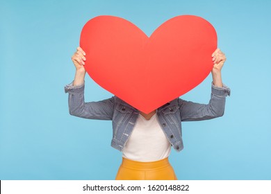 Anonymous Woman In Casual Outfit Hiding Face Behind Huge Red Heart, Concept Of Secret Love Fondness, Unknown Lover And Affair, Romance On Valentines Day. Indoor Studio Shot Isolated On Blue Background