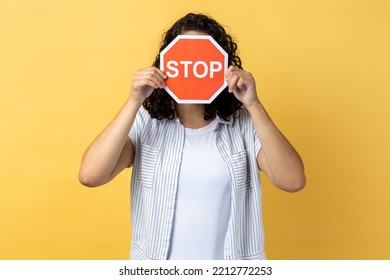 Anonymous Woman With Dark Wavy Hair Standing Covering Her Face With Red Stop Sign, Warning About Problems, Concept Of Ban, Denial, Prohibited To Go. Indoor Studio Shot Isolated On Yellow Background.