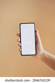 An Anonymous Woman Holding Mobile Phone With Empty Screen On Brown Background.  
Close Up Photo Of Female Hand Showing Smartphone With Blank Screen Isolated Over A Beige Background.