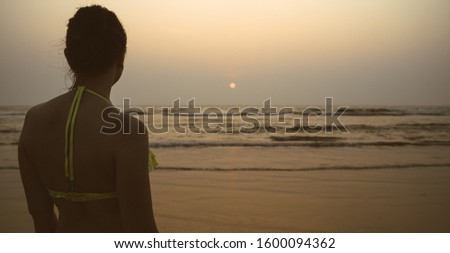 Anonymous woman near sea during sunset. Back view of unrecognizable female in swimwear standing on beach near waving sea in evening on resort