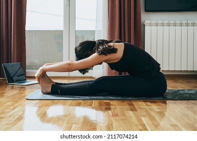 An Anonymous Woman Watching Training Videos On Her Laptop Computer At Home. 
Side View Of Fitness Woman In Sportswear Practicing Yoga On A Gray Mat And Using Laptop For Online Video Tutorials.