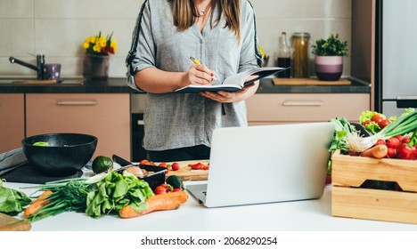 An Anonymous Woman Writing New Recipe In A Notebook And Watching Cooking Tutorial On A Laptop Computer While Standing At Table Full With Fresh Colorful Vegetables For Homemade Lunch 