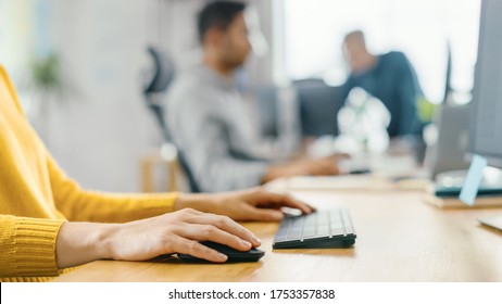 Anonymous Young Woman Sitting At Her Desk Using Laptop Computer. Focus On Hands Typing On Keyboard. In The Background Bright Office Where Diverse Team Of Young Professionals Work.