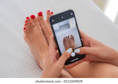 an anonymous young woman taking a photo of her feet with red painted nails while lying in a white bed relaxed. Natural light and real people - Powered by Shutterstock