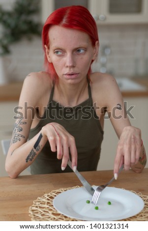 Anorexic woman. Skinny anorexic woman with tattoos holding knife and fork sitting near empty plate