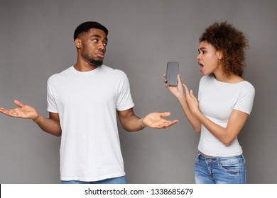 Another Life Online. Angry African-american Girlfriend Asking For Explanation To Her Cheater Boyfriend, Point At His Smartphone, Gray Studio Background