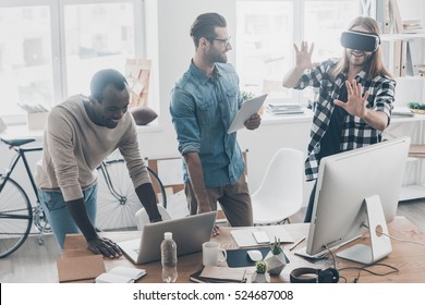 Another Reality Is Here! Group Of Young Business People Working And Communicating Together While One Of Them Wearing VR Headset In Creative Office