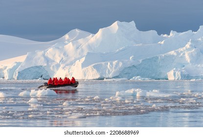 Antarctic expedition, cruise passengers in red parkas ride in a Zodiac inflatable boat, very close to a huge white iceberg in Cierva Cove bay - Shutterstock ID 2208688969