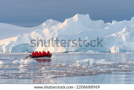 Antarctic expedition, cruise passengers in red parkas ride in a Zodiac inflatable boat, very close to a huge white iceberg in Cierva Cove bay